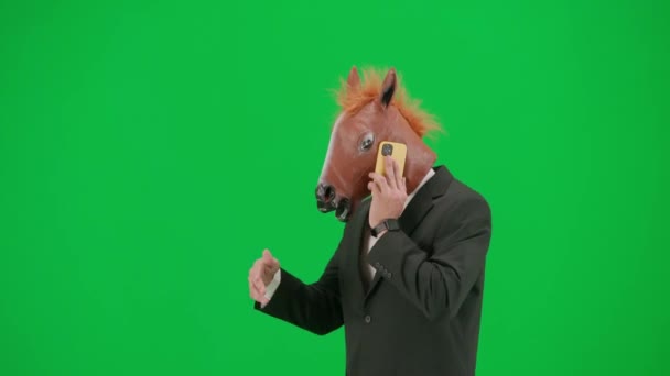 グリーンスタジオの背景に馬のヘッドマスクとビジネスススーツの男 スマートフォンで歩いて話すビジネスマン ハードオフィスのコンセプト ハーフターン — ストック動画
