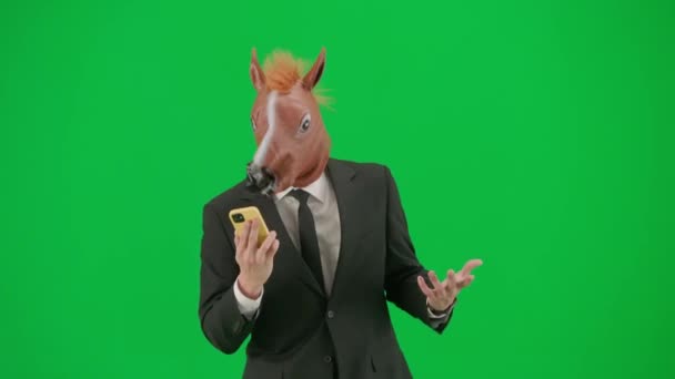 グリーンスタジオの背景に馬のヘッドマスクを持つビジネスススーツの男 ビジネスマンはスマートフォンで話し 怒って電話に叫びます ハードオフィスワークコンセプト — ストック動画