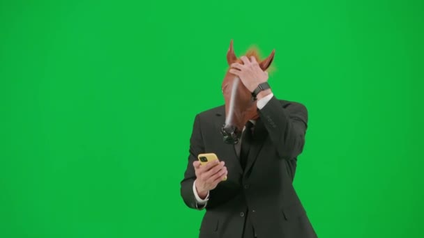 一个穿着西装 戴着马头面具 背景为绿色工作室的男人 一位商人正在看他智能手机上的某样东西 对失败感到很沮丧 艰苦工作概念 — 图库视频影像