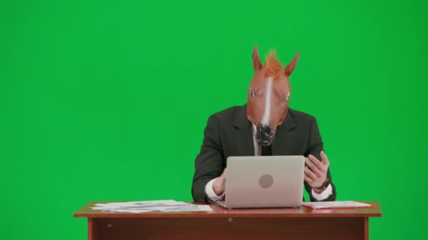 スタジオグリーンの背景に馬のヘッドマスクとビジネスススーツの男 ビジネスマンは ノートパソコンのキーボードにタイピングし 非常に怒っている机に座っています ヘビーオフィスワークコンセプト — ストック動画