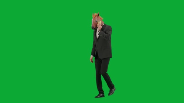 身穿西装 头戴马头面具 背景是绿色工作室 商人一边走路一边用智能手机说话 勤勤恳恳工作的概念 — 图库视频影像