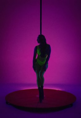 Sarı neon örgü elbiseli genç bir kadın direğin yanında poz veriyor. Yüksek topuklu dansçı stüdyoda pembe arka planda mavi ışıklı.