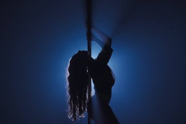 Esnek ve plastik kadın gölgesi direğin üzerinde dans ediyor. Uzun saçlı dansçı, karanlık stüdyoda spot ışıklarına karşı direkte poz veriyor.