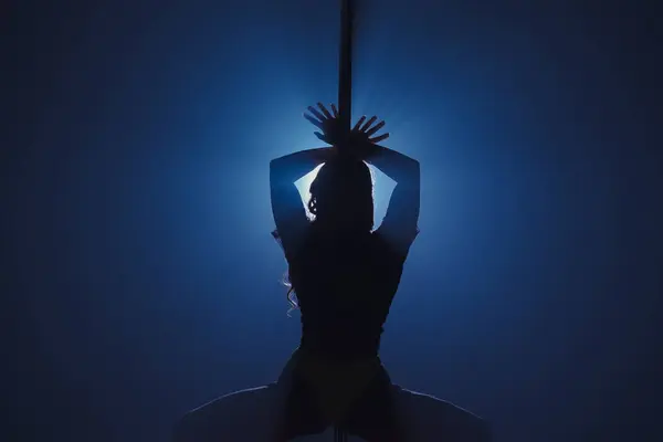 ポールで踊る柔軟でプラスチックな女性のダークシルエット 明るいスポットライトに対して暗いスタジオでピロンの長い髪のダンサー — ストック写真