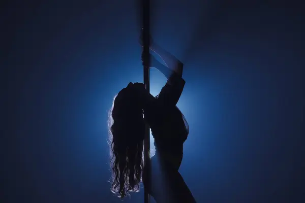 ポールで踊る柔軟でプラスチックな女性のダークシルエット 明るいスポットライトに対して暗いスタジオでピロンの長い髪のダンサー — ストック写真