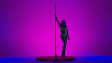 Genç muhteşem kadın direk dansı sırasında esnekliğini ve esnekliğini gösteriyor. Sarı takım elbiseli kadın stüdyoda direkte dans ediyor. Pembe arka planda, mavi ışıklı.