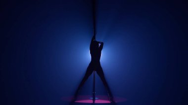 Esnek ve plastik bir kadının karanlık silueti direğin üzerinde dans ediyor. Karanlık bir stüdyoda, parlak bir ışığa karşı direkte dans eden bir kadın.