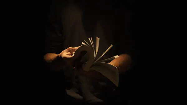 Historical and vintage objects creative advertisement concept. Studio shot of old retro hardcover book on dark background in warm light. Man holding flipping through pages of old book in hands.