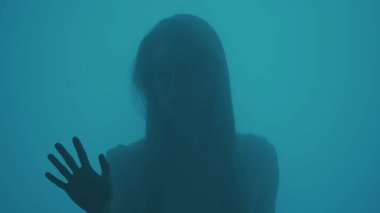 Güzellik ve kozmetoloji yaratıcı reklam konsepti. Buhar ve su damlaları içinde cam pencerenin arkasındaki mavi neonlu kadın portresi. Camın önünde poz veren makyajlı ve saç stili olan kız..