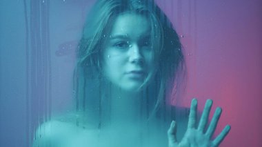 Güzellik ve kozmetoloji yaratıcı reklam konsepti. Cam pencerenin arkasındaki neon ışıklı kadın portresi. Buhar ve su damlaları. Çekici kız cama tutunup bakıyor..