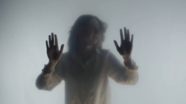 Şeffaf bir buzlu perde ya da camın arkasında melankolik bir kadın silueti. Bir kadın avuçlarıyla perdenin yüzeyine dokunuyor.