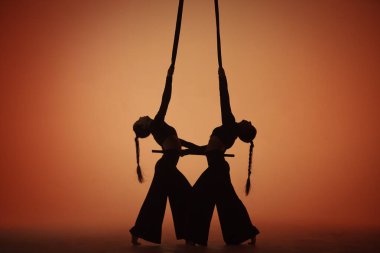 Stüdyonun turuncu arka planında iki akrobatın karanlık siluetleri hava trapezinin yanında duruyor. Hava jimnastikçileri ayna görüntüsü gibi. Sirk gösterisi