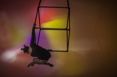 Stüdyonun çok renkli arka planında siyah taytlı hava jimnastikçisi. Kadın akrobat bir küp üzerinde, akrobatik koreografinin elementlerini sergiliyor.