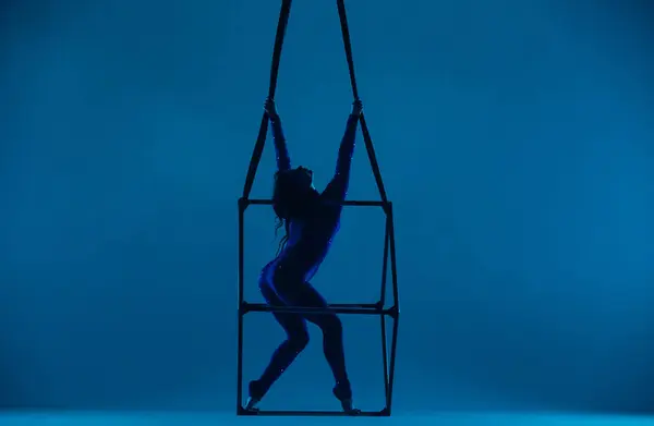Silhouette of female aerial gymnast isolated on blue neon background. Female acrobat performing in the air on a cube, showing her flexibility