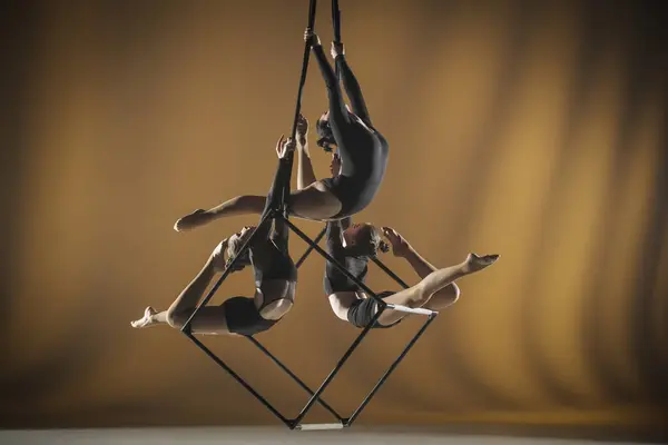 Trio of young female acrobats posing on a cube suspended at a height. Aerial gymnasts perform in a studio with yellow lights and demonstrate flexibility and stretching. The concept of a circus show