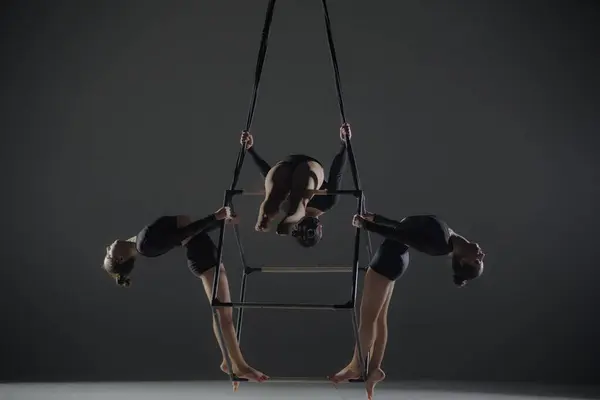 Trio of young female acrobats posing on a cube suspended at a height. Aerial gymnasts perform in studio against dark background and demonstrate flexibility and stretching. The concept of a circus show