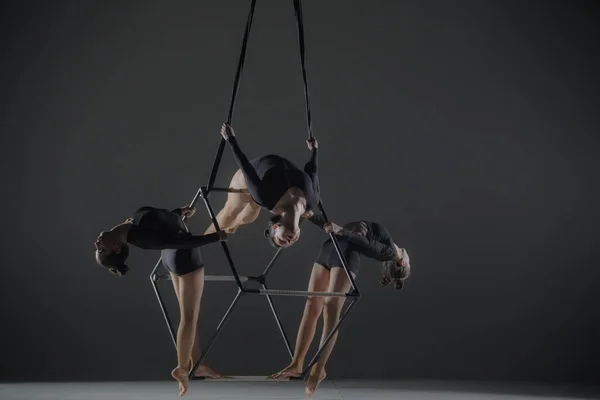 Trio of young female acrobats posing on a cube suspended at a height. Aerial gymnasts perform in studio against dark background and demonstrate flexibility and stretching. The concept of a circus show