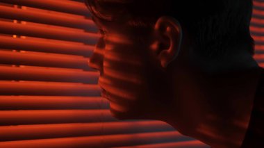Siluet soyut yaratıcı reklam konsepti. Karanlık odadaki erkek portresi. Pencerenin yanında yakışıklı bir adam, kırmızı neon ışıkları jaluzinin arkasında parlıyor, adam dışarıya bakıyor, aşırı yakın çekim..