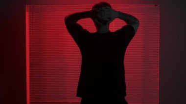 Silüet estetik yaratıcı reklam konsepti. Karanlık stüdyoda erkek modelin portresi. Tişörtlü genç adam pencerenin önünde kırmızı ışıkta duruyor ve jaluzinin arkasında ellerini kafaya koyuyor..