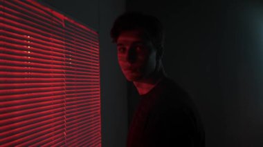 Silüet estetik yaratıcı reklam konsepti. Karanlık stüdyoda erkek modelin portresi. Tişörtlü genç adam pencerenin yanında duruyor. Kırmızı ışık arkamızda. Jalousie kameraya arkasını dönüyor..