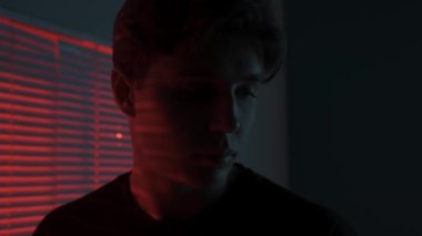 Silüet estetik yaratıcı reklam konsepti. Karanlık stüdyoda erkek modelin portresi. Pencere kenarında duran tişörtlü genç adam, arkasında kırmızı ışık, akıllı telefondan sesleniyor..