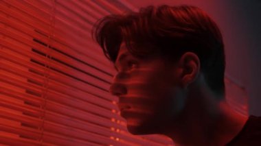 Silüet estetik yaratıcı reklam konsepti. Karanlık stüdyoda erkek modelin portresi. Pencere kenarında duran tişörtlü genç adam, arkasında kırmızı ışık olan Jalousie pencereden dışarı bakıyor..