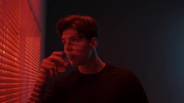 Silüet estetik yaratıcı reklam konsepti. Karanlık stüdyoda erkek modelin portresi. Tişörtlü genç adam pencerenin yanında kırmızı ışıkta duruyor. Jalousie 'nin arkasında içki içiyor. Dışarıya bakıyor..
