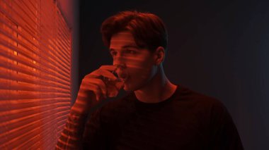 Siluet soyut yaratıcı reklam konsepti. Karanlık odadaki erkek portresi. Pencerenin yanında yakışıklı bir adam, kırmızı neon ışıkları külüstürün arkasında parlıyor, adam dışarıya bakıyor bardaktan viski içiyor..