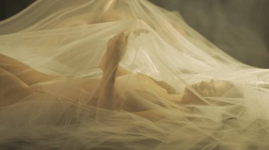 Yatakta, şeffaf bir hava bezinin altında genç bir kadının yan görüntüsü. Yaklaş. Sıcak ışıkla aydınlatılan bir kadın kollarını kaldırdı ve bir tül oluşturdu.