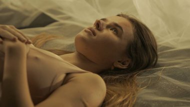 Kolları göğsünde katlanmış beyaz bir örgü kumaşın üzerinde hareketsiz yatan çekici bir kadın. Sıcak ışıkta beyaz örgü kumaşında gözleri açık bir kadın yatıyor.