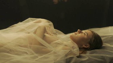 Kolları göğsünde bir aynanın yanında katlanmış, sırtüstü uzanmış bir kadının yan görüntüsü. Kadın boynuna kadar bembeyaz şeffaf bir tül bezle örtülmüş.