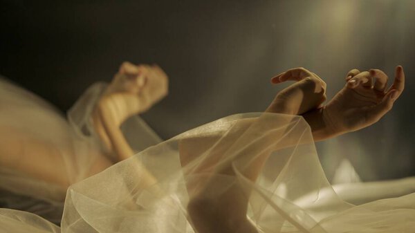 Hands of a woman with white tulle on a black background close up. A mirror reflects the blurred silhouette of a woman covered with netted fabric, illuminated by warm rays of light