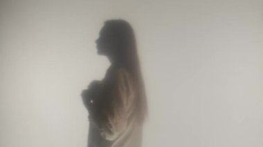 Beyaz bluzun düğmelerini açan sarışın kadının siluetinin yan görüntüsü. Buzlu camların ya da perdelerin arkasındaki buharlı ya da sisli bir kadının profil portresi HDR BT2020 HLG materyalini kapat