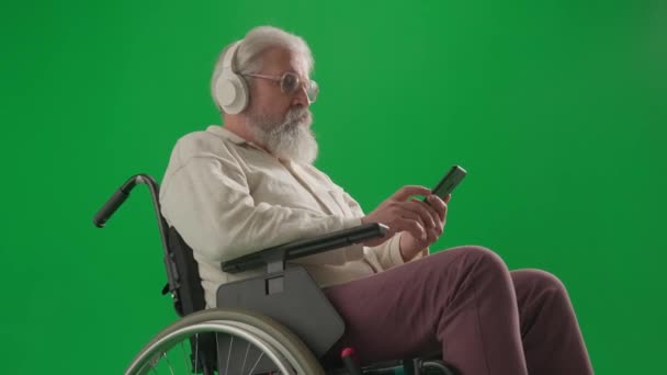年金受給と日常生活クリエイティブ広告コンセプト クロマ グリーン スクリーンの老化した障害者の肖像画 スマートフォンを持っているヘッドフォンの車椅子のシニア男性を閉じる — ストック動画