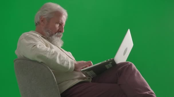 年金レジャーと日常生活クリエイティブ広告コンセプト クロマキーグリーンスクリーンの高齢のひげの男の肖像画 椅子に座ってラップトップで作業するカジュアルな服のシニア男性 — ストック動画