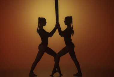 Akrobatik yaratıcı konsept. İki kadın akrobatın silueti sarı neon arka planda, merkezinde spot ışığı var. Kadın akrobatlar ellerinde havadan iplerle poz veriyorlar.