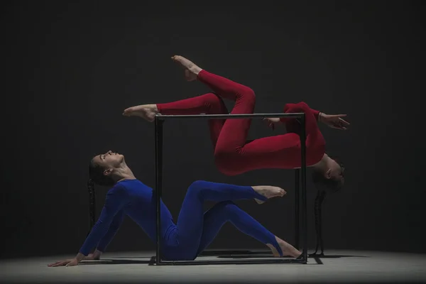 Modern choreography and acrobatics creative advertisement concept. Portrait of two female acrobats isolated on black background. Girls aerial dancers in blue red suits showing element on a cube.