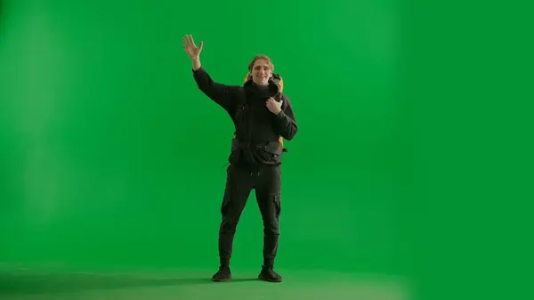 A walking male tourist with a hiking backpack on his back, stops and waves his hand in greeting when he sees his acquaintances. A man hiker in full height in the studio on the green screen