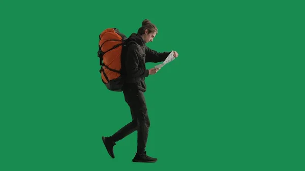 A male tourist studies a map of the area while hiking. A man traveler in full length with a backpack on his back walking in the studio on the green screen
