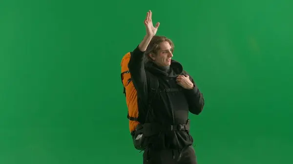 A walking male tourist with a hiking backpack on his back, stops and waves his hand in greeting when he sees his acquaintances. A man hiker in the studio on the green screen