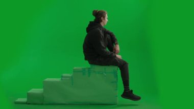Stüdyoda yeşil ekranda yüksek bir pozisyonda oturan bir erkek yolcunun yan görüntüsü. Adam bacakları havada oturur, elini gözlerine koyar ve uzaklığa bakar.