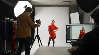 Fotoğraf üretimi ve yaratıcı takım çalışması konsepti. Model ve profesyonel takımın sahne arkası stüdyoda. Çekici kadın fotoğrafçı için poz veriyor, erkek editör masada oturmuş monitöre bakıyor..