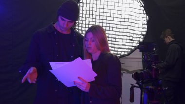 Kadın bir yapımcı ve videograf çekimlerden önce film tahtasını tartışırlar. Bu arada ışıklandırma teknisyeni ışıklandırma ekipmanlarını ayarlıyor. Video çekiminin perde arkası