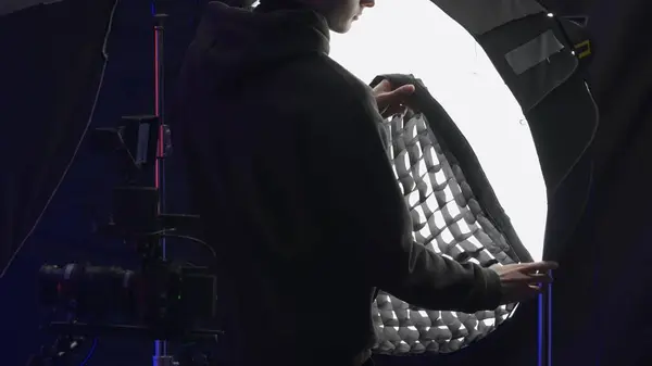Back view of a lighting technician putting a net on a spotlight to diffuse light, close up. Video recording studio with equipment, backstage