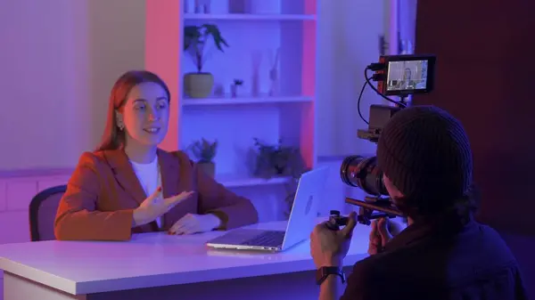 Backstage video production recording. A videographer uses a professional camera to film a female presenter sitting at a table in front of a laptop. Film crew in the studio in pink and blue neon