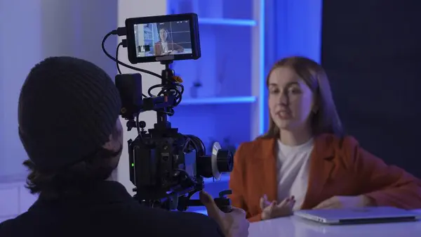 后台视频制作录音 摄影师用专业相机拍摄坐在笔记本电脑前桌子旁的女主持人 电影摄制组在蓝色霓虹灯下拍摄 — 图库照片