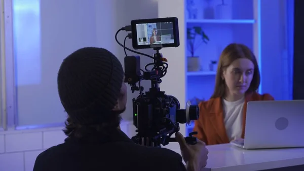 Backstage video production recording. A videographer uses a professional camera to film a female presenter sitting at a table in front of a laptop. Film crew in the studio in blue neon lighting