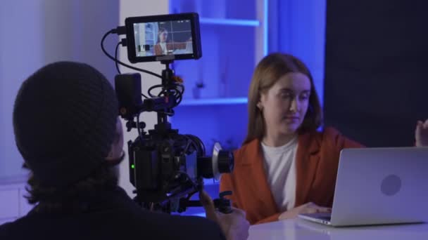 バックステージビデオ制作記録 ビデオグラファーはプロのカメラを使って ラップトップの前にテーブルに座っている女性のプレゼンターを撮影します ブルーネオン照明のスタジオの映画クルー — ストック動画
