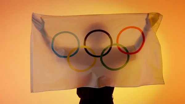 Sports flags advertisement concept. Person silhouette holding big flag with five rings against yellow background. Silhouette of man with national Olympic logo flag holding in hands.