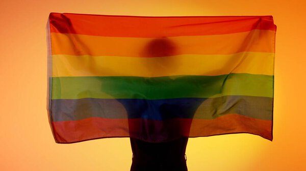Love and diversity advertisement concept. Person silhouette holding rainbow flag against yellow background. Silhouette of man with LGBT pride flag holding in hands.